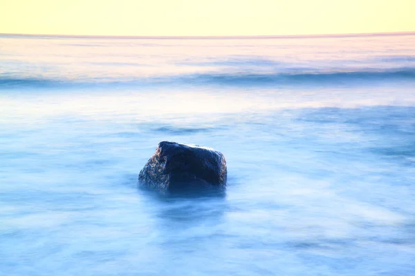 海の静かな朝でロマンチックな雰囲気。大きな岩が滑らかな波状の海から突き出ています。ピンクの地平線 — ストック写真