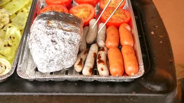 Grilling white bratwurst and red sausages, between tomatoes, lump in alu foil and white peppers in AL case on gas barbeque grill — Stock Video