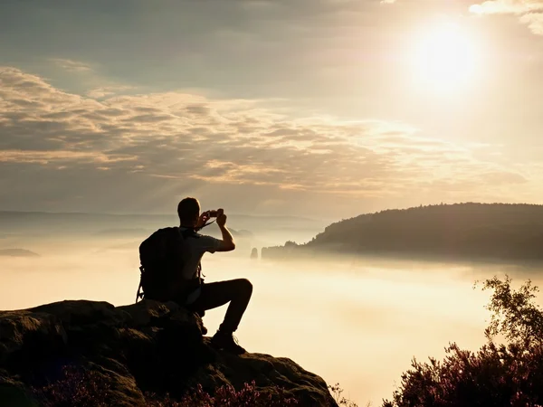 Dreamy fogy landscape. Amateur photographer takes impressive photos with phone. Hiker with backpack on cliff photogrphy  orange pink misty sunrise in a beautiful valley below — Stock Photo, Image