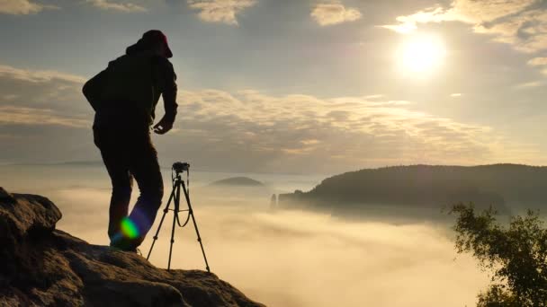 Fotograf w zielonej wiatrówka i czarne Spodnie trekkingowe zatrzymać aparat na statywie na krawędzi klifu. Dreamy fogy krajobraz, niebieski mglisty wschód słońca w pięknej dolinie poniżej — Wideo stockowe