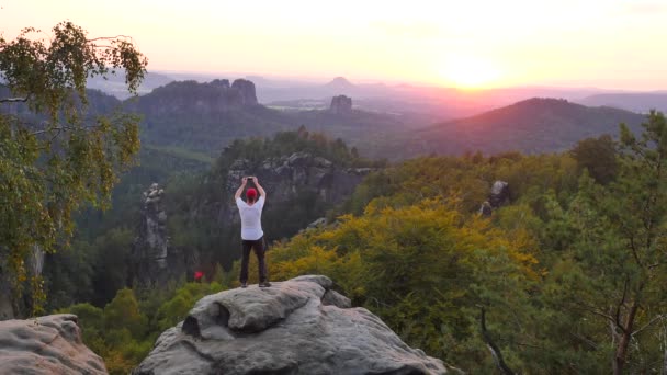 Turista en camisa gris y pantalón de trekking fotografía. El hombre toma fotos o graba video con un teléfono inteligente en el pico del parque del imperio del rock. Atardecer de verano sobre el hermoso valle del parque Sajonia Suiza . — Vídeos de Stock