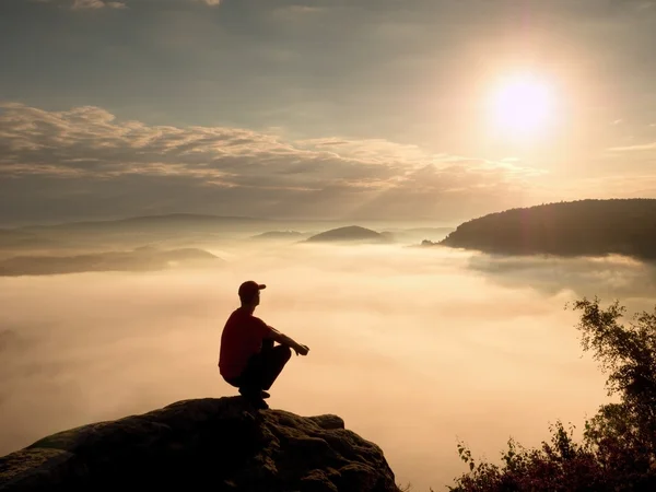 Man tourist  sit on rock empire. View point with heather and branches  above misty valley. Sunny daybreak in rocky mountains. — Stock Photo, Image