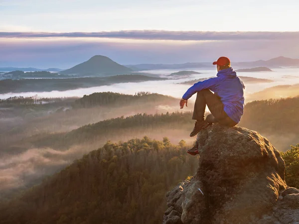 Misty morning in pure nature. Sitting man on sharp rocky edge, enjoy amazing birds view.  Misty fall landscape bellow.