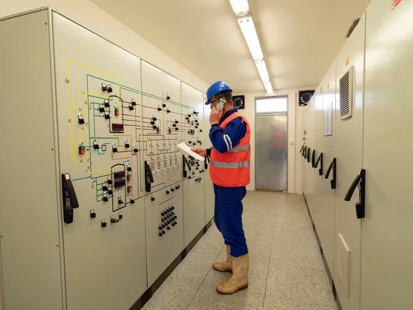 労働者は大きな制御室にいる スキーマを使用した発電所管理パネル コントロールパネルの前に立つエンジニア監視室と値と電話を確認してください — ストック写真