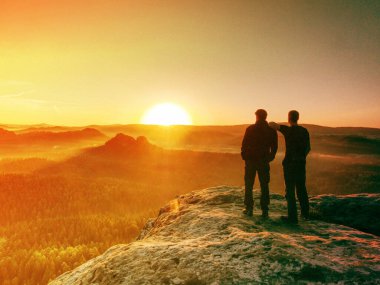 Two tourists, men friends standing together and looking over misty mountain landscape with coniferous forest.  Abstract filter. clipart