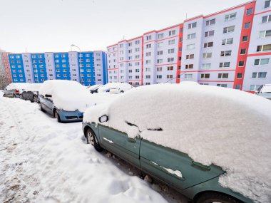 Terk edilmiş arabalar kışın bir kar yığınının altında otoparkta gömülüdür. Arka planda renkli evler