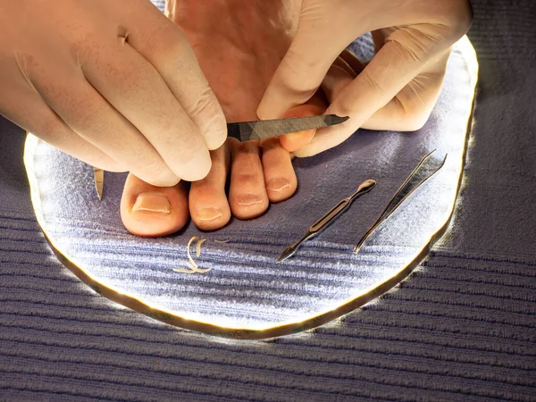 Hands in white sterile gloves file toenails with long pointed file on male toes. Pedicure on blue towel