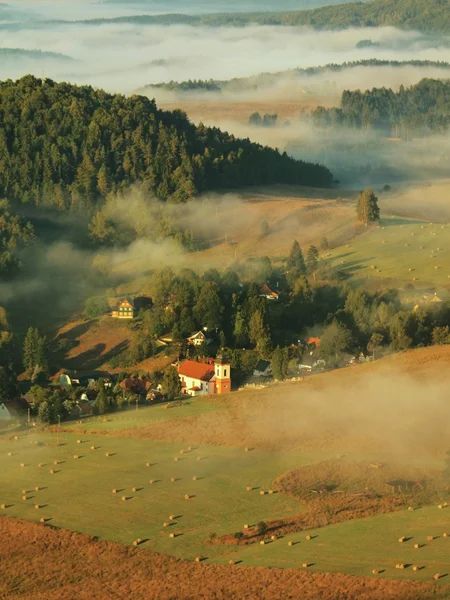 Alba soleggiata in una splendida campagna del parco della Svizzera ceco-sassone. Delicata nebbia sopra la chiesa del villaggio. I raggi del sole caldi combattono con nebbia di terra fredda in valle . — Foto Stock