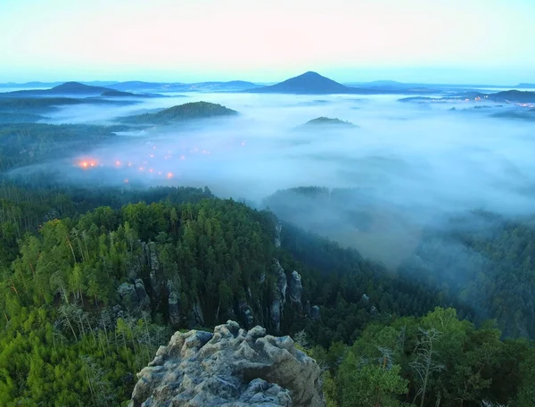 Valle brumoso azul después de la noche lluviosa, luces de color naranja rosado están brillando desde el pueblo de abajo. La niebla se mueve entre colinas y picos de árboles, cielo azul . — Foto de Stock