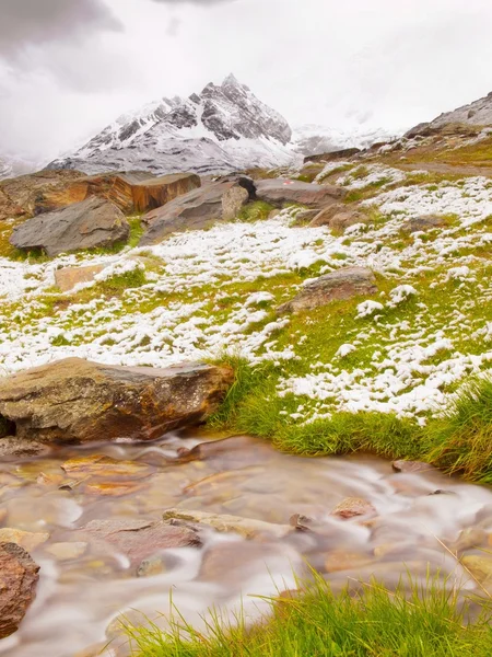 Första snön på alpina äng. snabb ström faller ner över toffel stenar till djupt misty valley. snöiga toppar i Alperna bergen i bakgrunden. — Stockfoto