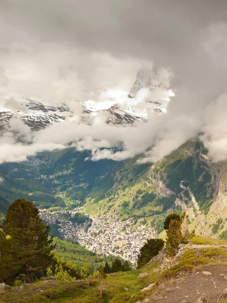 Diepe vallei hieronder nevelige pieken van de Alpen bergen in de achtergrond. kleine berg stad onder dekking van de nevel. — Stockfoto