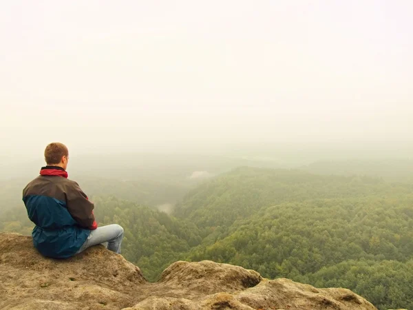 Homem triste sentar-se no pico da pedra de arenito e olhando sobre o nevoeiro e nebuloso vale da manhã . — Fotografia de Stock