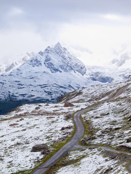 Paysage enneigé avec route graveleuse. Sommets pointus et brumeux de hautes montagnes en arrière-plan . — Photo
