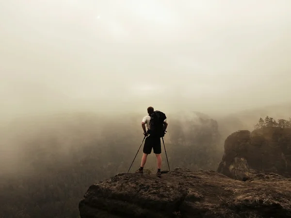 Turista com postes e mochila grande em pé no ponto de vista rochoso e observando a paisagem nebulosa da manhã. Parque nacional Saxon Suíça na Alemanha. manhã de outono melancólica . — Fotografia de Stock