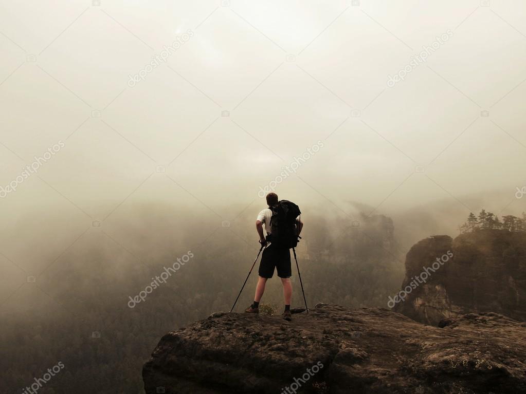 Tourist with poles and big backpack standing on rocky view point and watching into misty morning landscape. National park Saxon Switzerland in Germany. Melancholic autumn morning.
