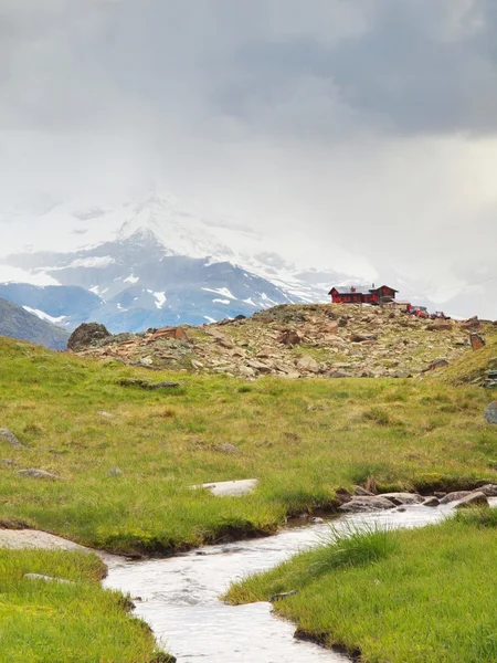 Bach in frischen grünen Almwiesen, schneebedeckte Gipfel der Alpen im Hintergrund. Kaltes nebliges und regnerisches Wetter in den Bergen zum Ende des Sommers — Stockfoto