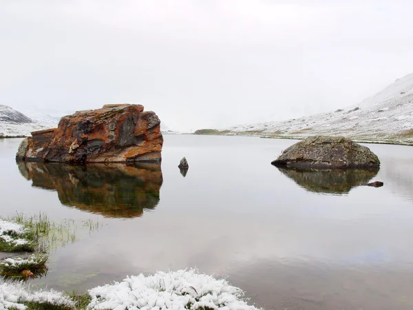 Der erste Schnee am Alpensee. Herbstsee in den Alpen mit Spiegelebene und schneebedecktem Gras und Geröll ringsum. neblig scharfe Gipfel hoher Berge im Hintergrund. — Stockfoto