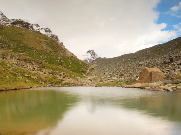 Autumn alpine lake with mirror level. Stony hills above lake banks and sharp snowy and misty peaks of high mountains. Dark cloudy sky. — Stock Photo, Image