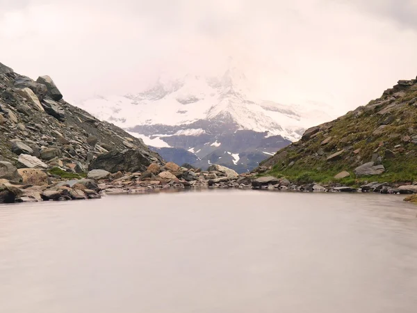 Höstens alpine lake med spegel. Steniga bergen ovanför sjön banker och skarpa snöiga och dimmiga toppar av höga berg. Mörka molnig himmel. — Stockfoto