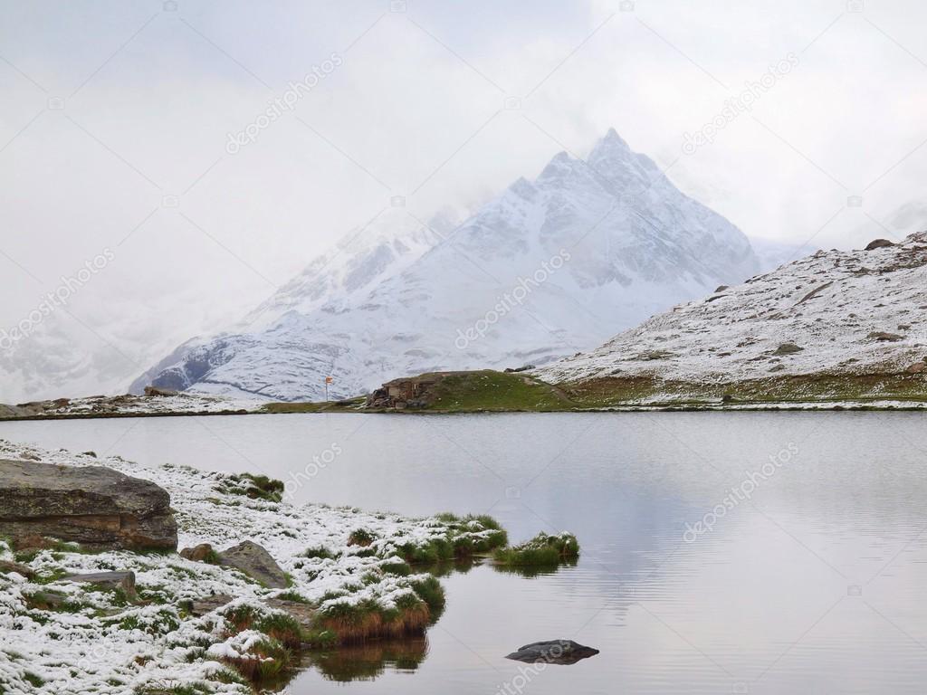 First snow at Alpine lake. Autumn lake in Alps with mirror level and snowy grass and boulders around. Misty sharp peaks of high mountains in background.