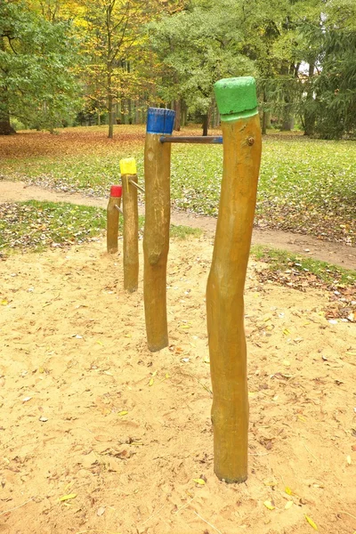 Ocelová hrazda na dřevěných sloupech v dětské hřiště. oranžový písek pod pruhy, zelený park v pozadí. — Stock fotografie