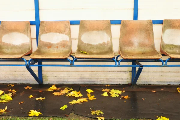 Alte Plastiksitze auf der Spielerbank im Freien, Stühle mit abgenutzter Farbe unter dem gelben Dach. Herbstlaub, Ende der Fußballsaison. — Stockfoto