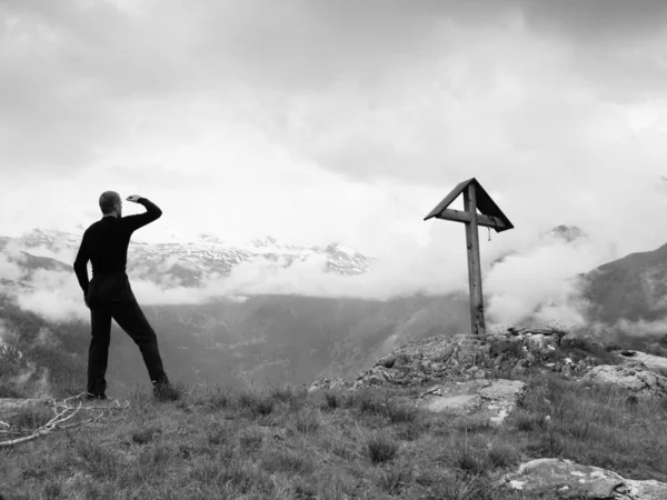 Touristen stehen auf felsigen Aussichtspunkten und blicken ins neblige Alpental. Holzkreuz auf einem für die Alpen typischen Berggipfel. Schwarz-Weiß-Foto. — Stockfoto