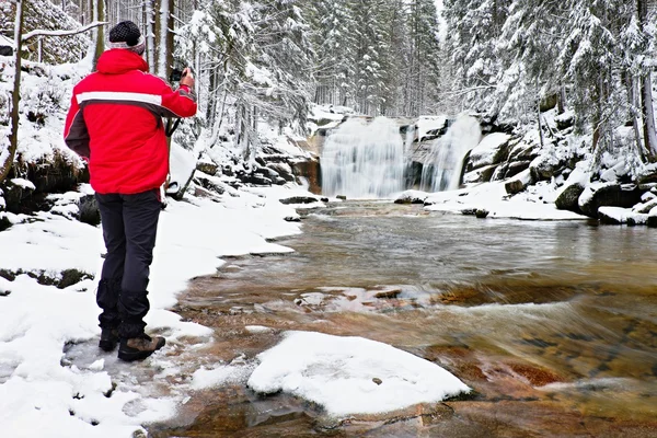 Fotografia em casaco vermelho com câmera digital nas mãos está tirando foto da cachoeira de inverno — Fotografia de Stock