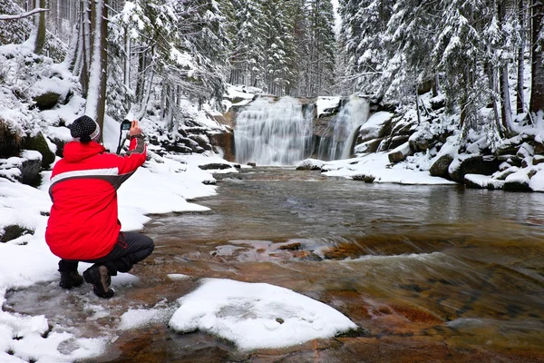 Fotografia em casaco vermelho com câmera digital nas mãos está tirando foto da cachoeira de inverno — Fotografia de Stock