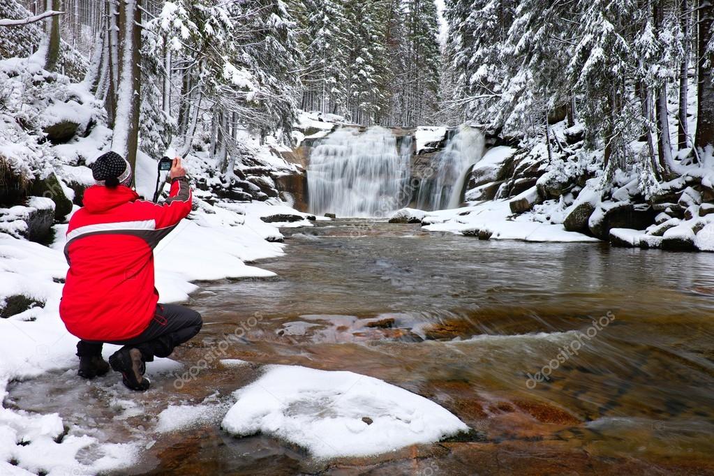 Photograph in red jacket with digital camera in hands is taking photo of winter waterfall