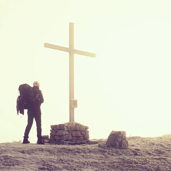 Tourist with big backpack stand at cross memorial on mountain peak. Man is watching into misty Alpine valley bellow. Metal cross at a mountain peak. — Stock Photo, Image