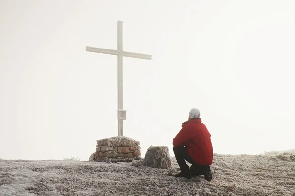 Tourist in red is kneeling  at cross memorial on mountain peak. Man is watching into misty Alpine valley bellow. Metal cross at a mountain peak. — Stock Photo, Image