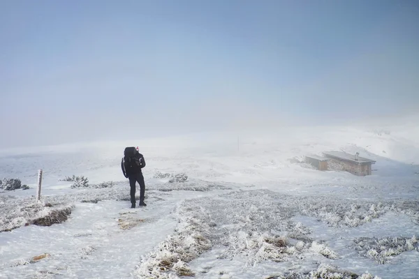 Samotné turistické s velkým batohem a sněžnice chodit na zasněžené cestě do mlhy. Národní park Alpy park v Itálii. Pogy zimní počasí. Malý domek v pozadí. — Stock fotografie