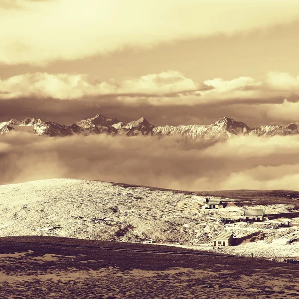 Huts at peaks of Alps hill, sharp rocky mountains at horizon. Sunny winter day. Frozen stalk of grass in large meadow with powder snow. — Stock Photo, Image