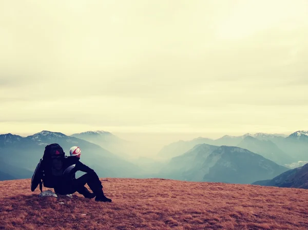 Tired out tourist in black with backpack is sitting on stone in meadow and watching into misty valley. Autumn misty day in mountains. — Stock Photo, Image