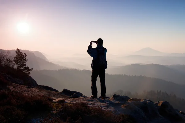 L'uomo scatta foto con smart phone sulla vetta dell'impero rock. Paesaggio nebbioso da sogno, primavera arancio rosa nebbia alba in una splendida valle del parco della Sassonia Svizzera . — Foto Stock