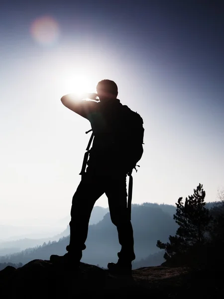 Турист зі спортивним рюкзаком стоїть на скелястій точці зору і дивиться в туманну долину. Сонячний туманний ранковий спалах у скелястих горах . — стокове фото