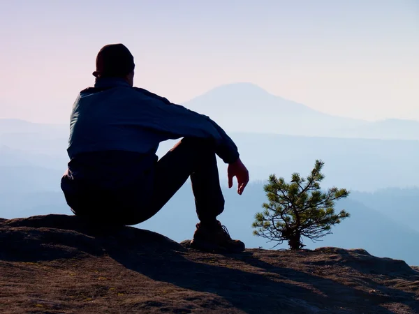 Турист в синей ветровой куртке сидит на скалистой смотровой площадке и наблюдает за туманной долиной. Солнечный туманный рассвет в скальных горах . — стоковое фото