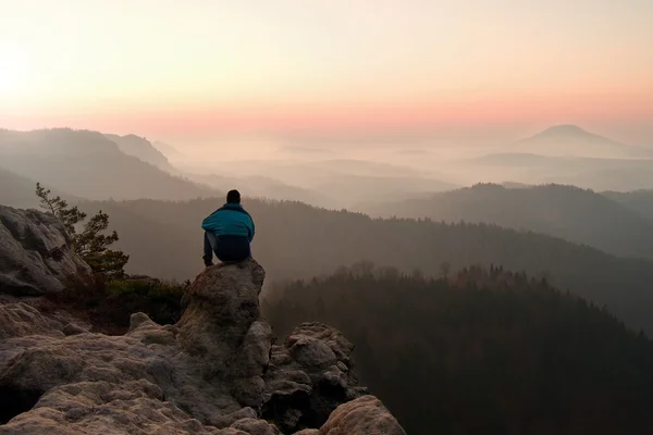 Turista en chaqueta de viento azul sentarse en el punto de vista rocoso y ver en el valle brumoso a continuación. Soleado amanecer brumoso en las montañas rocosas . —  Fotos de Stock