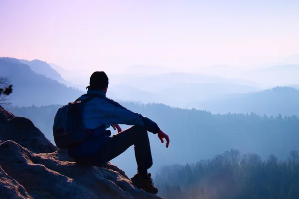 Turysta z sportowy plecak siedzieć na skalisty szczyt i oglądanie w mgliste doliny poniżej. Słoneczny świt zamglony w Górach Skalistych. — Zdjęcie stockowe