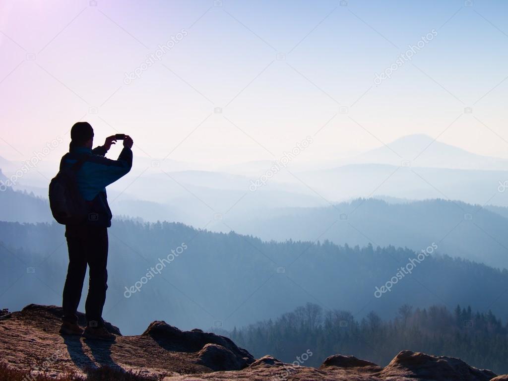 Tourist takes photos with smart phone on peak of rock. Dreamy fogy landscape, spring orange pink misty sunrise in a beautiful valley below rocky mountains.