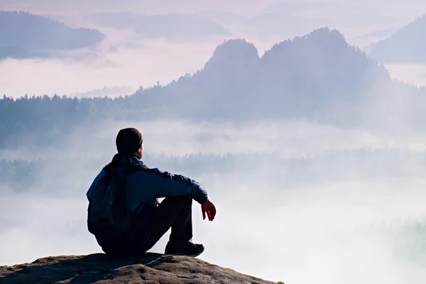 Tired tourist in windcheater and dark cap sit on rock and watching into blue misty valley bellow. Sunny spring daybreak in rocky mountains. — Stock Photo, Image