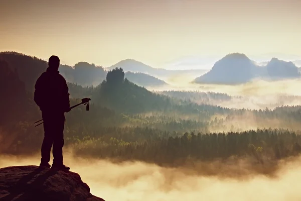 Alto zaino in spalla con pali in mano. Sunny alba di primavera in montagne rocciose. Escursionista con zaino in piedi su un punto di vista roccioso sopra la valle nebbiosa . — Foto Stock