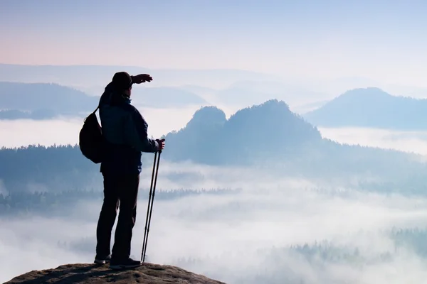 Tall backpacker with poles in hand. Sunny spring daybreak in rocky mountains. Hiker with backpack stand on rocky view point above misty valley.