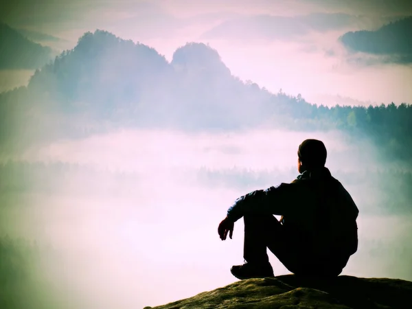Уставший турист в обманщике и темной шапке сидит на скале и наблюдает за голубой туманной долиной. Солнечный весенний рассвет в скалистых горах . — стоковое фото
