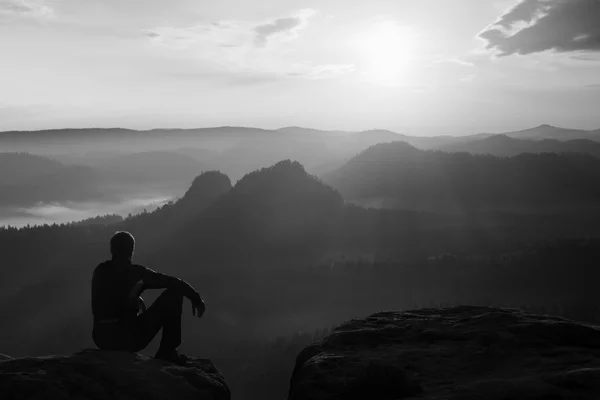 Touristen in schwarzer Windjacke sitzen auf einem scharfen Felsen und blicken in ein buntes Nebeltal. Sonnenaufgang im Frühling in den felsigen Bergen. — Stockfoto
