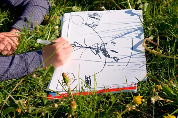 Vista de perto da mão da criança que desenha ornamentos coloridos no papel. Caderno na grama verde fresca do prado . — Fotografia de Stock