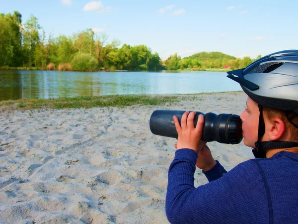 Niño pequeño con un casco de bicicleta en la cabeza, en ropa deportiva tiene una botella de plástico de agua en la mano y bebidas . — Foto de Stock