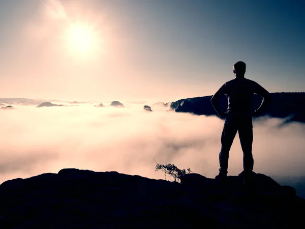 Homem de camisa e jeans está de pé no pico do império arenito e olhando sobre o nebuloso e nebuloso vale da manhã para o sol — Fotografia de Stock