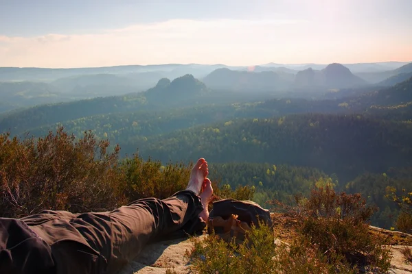 Pernas masculinas cansadas em calças de caminhada escuras descansam no pico da rocha acima do vale . — Fotografia de Stock